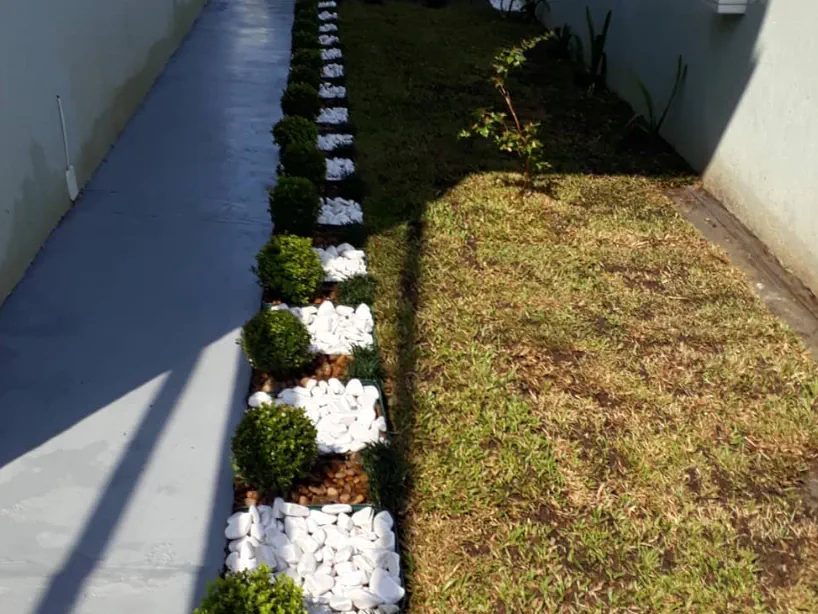 Canteiro lateral com buxinhos, pedras brancas e gramado após projeto de paisagismo